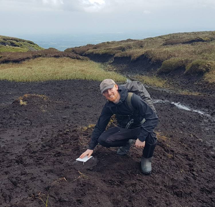 Kieran is crouching on a degraded area of blanket bog. He is wearing a faded ball cap, black rain coat, trousers and rain boots. Kieran is smiling at the camera and his hand is resting on a pitfall trap.