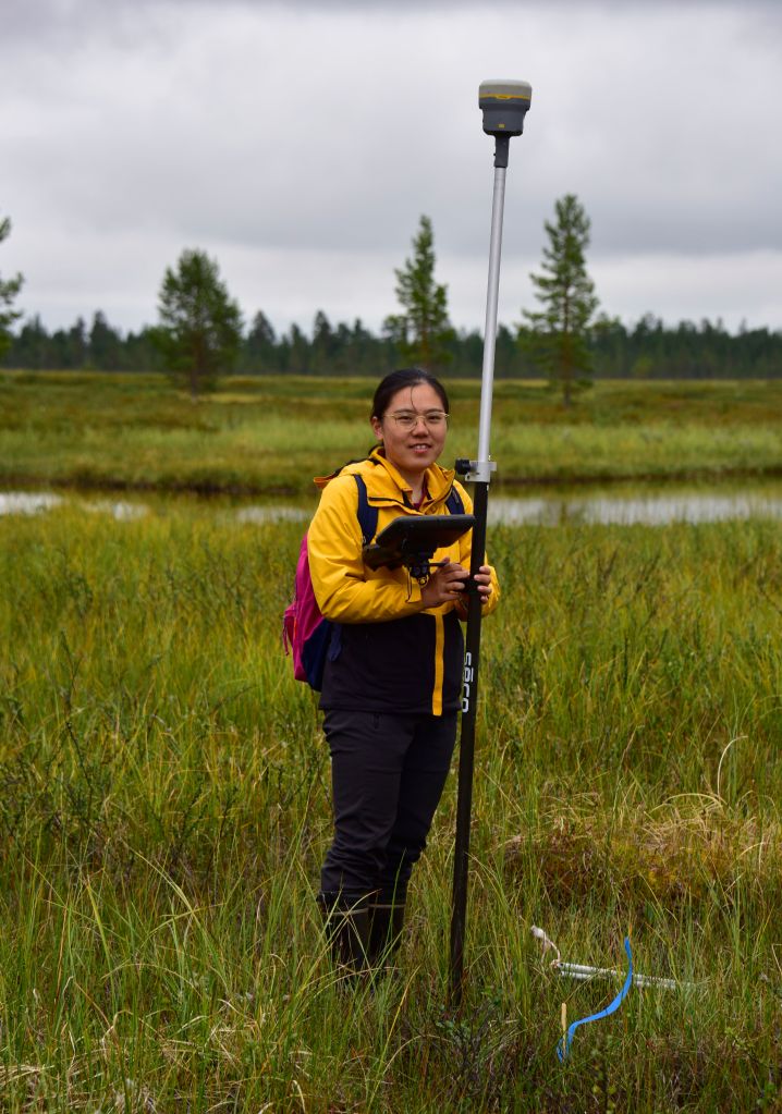 Yuwen is standing in a peatland wearing a yellow and black coat, black trousers, and pink backpack. Yuwen is smiling at the camera and holding a differential GPS.